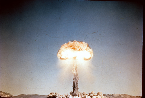Vintage 35mm slide scan of a historic atom bomb explosion and mushroom cloud exploding in the middle of the desert. Mountain range on the horizon. Center of mushroom cloud is white-hot. Scratches on film. Dust cloud on the ground and tall mushroom cloud glowing and raining down