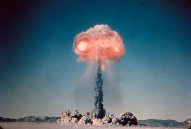 Old slide scan of atom bomb exploding in the desert with red hot fire cloud at the top Vintage 35mm slide scan of a historic atom bomb explosion and mushroom cloud exploding in the middle of the desert. Mountain range on the horizon. Scratches on film. Dust cloud on the ground and tall mushroom cloud glowing and raining down nuclear fallout stock pictures, royalty-free photos & images