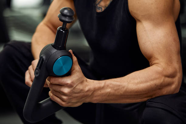 A young man is using and enjoying percussion massage gun to relax and relief the pain in body and muscles in the gym stock photo