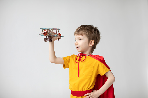 Dreamy boy in a red cape plays with an airplane toy. Little super hero.