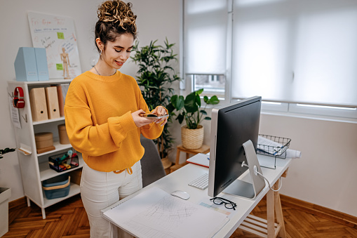 Young businesswoman wearing yellow sweatshirt photographing document with smart phone on desk by computer in office
