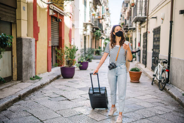 istock Young woman wearing protective face mask pulls suitcase down cobblestone street 1369213255