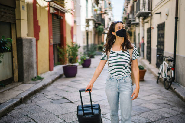 istock Young woman wearing protective face mask pulls suitcase down cobblestone street 1369213326