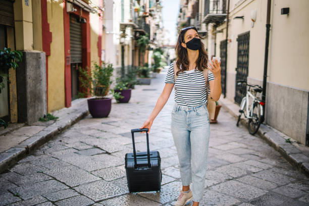 istock Young woman wearing protective face mask pulls suitcase down cobblestone street 1369213858