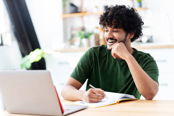 istock Indian young man using laptop for watching webinar, studying online and takes notes in a notebook, male student is studying online, e-learning concept 1369214157