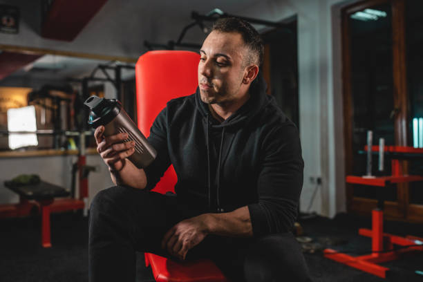 Front view of adult man one caucasian male athlete sitting on the bench at gym holding dark black supplement shaker while taking a brake in training bodybuilding weight lifting copy space vista frontal de un hombre adulto un atleta masculino caucásico sentado en el banco del gimnasio sosteniendo un agitador de suplementos negro oscuro mientras frena en el entrenamiento de culturismo levantamiento de pesas espacio de copia