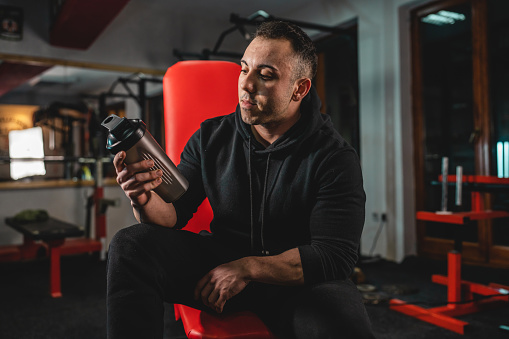 Front view of adult man one caucasian male athlete sitting on the bench at gym holding dark black supplement shaker while taking a brake in training bodybuilding weight lifting copy space