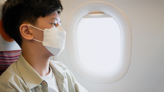 Handsome Asian teenager boy with face mask traveling on airplane, sitting and looking out the window.