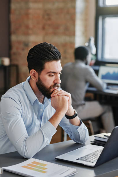 istock Vertical medium shot of young Middle Eastern man working on office sitting at desk looking at computer screen 1369248766