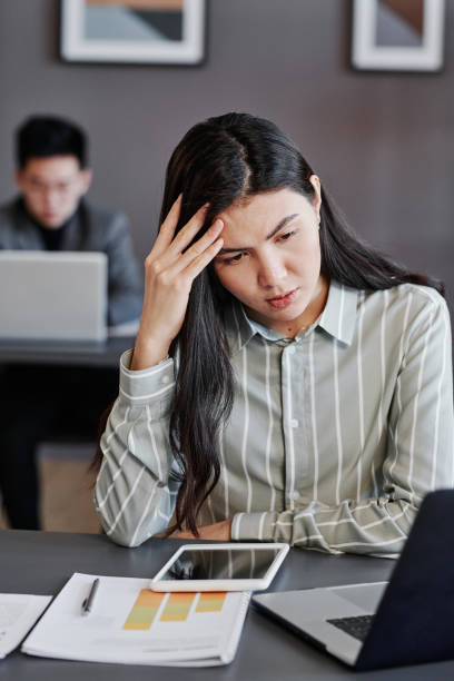 istock Vertical medium portrait of young Asian woman working on difficult data in office rubbing her head 1369251590