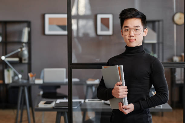 istock Horizontal medium portrait of handsome young Asian man working in modern office holding papers looking at camera 1369254082