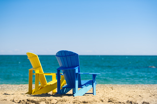 Two Adirondack chairs on a sandy beach facing a blue calm lake. The chairs are blue and yellow.