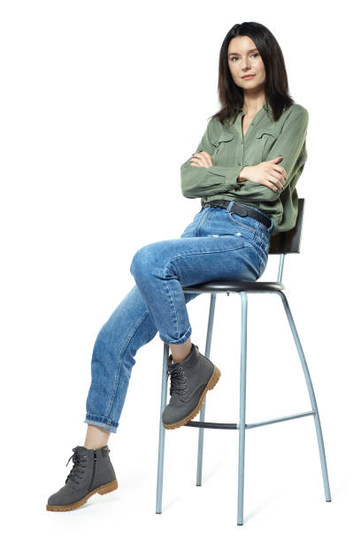 A young woman in jeans, boots and a khaki shirt is sitting on a high chair. Isolated on white. stock photo