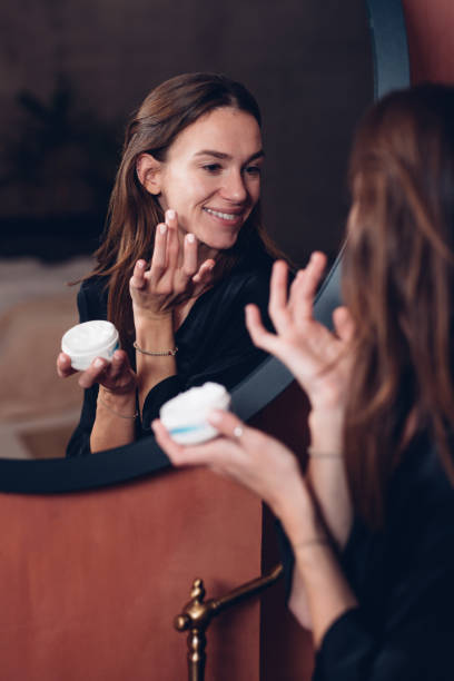 Young woman in front of the mirror putting cream on her face stock photo