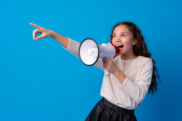 istock Cute schoolgirl holding megaphone against blue background 1369268902