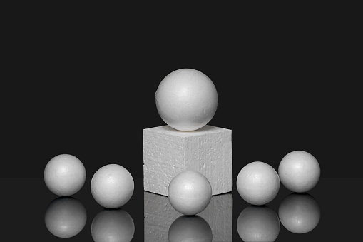 White polystyrene ball on 3d square podium, surrounded by small spheres. Black isolated background with reflection from objects. Geometric composition of various shapes.