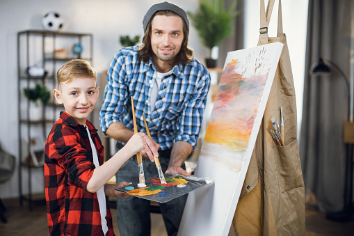 Portrait of handsome caucasian man and cute little boy looking at camera while holding paintbrush and color palette in hands. Father and son drawing together on easel at home.