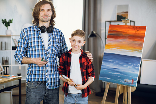 Caucasian young father embracing his cute son while posing at bright living room with paint brushes in hands. Colorful drawing standing on background.