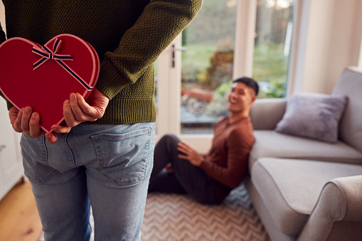 Man Surprising Same Sex Partner At Home With Heart Shaped Gift Held Behind His Back