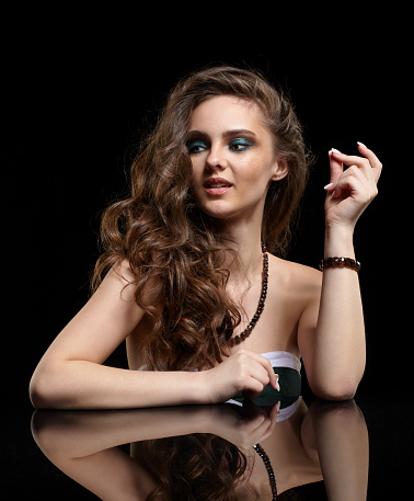 Beauty portrait of young woman sit at the black table. Brunette girl with long hair and evening female makeup on black background.