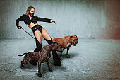 Beautiful and sexy blonde woman with slick back hair, wearing a leather muzzle, holding back an agressive dog while standing beside another one, ordering to attack while pointing in front of her