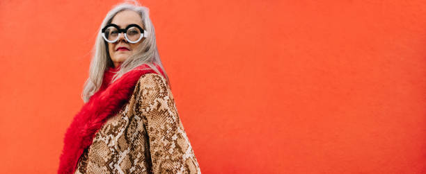 Grey-haired woman looking thoughtful against a red wall Grey-haired woman looking thoughtful while standing against a red background. Pensive elderly woman reflecting on memories of the past. Stylish senior woman wearing eyeglasses and colourful clothing. eccentric stock pictures, royalty-free photos & images