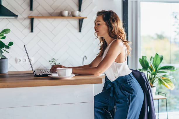 istock Serious woman sitting at table and typing on laptop in kitchen 1369301994