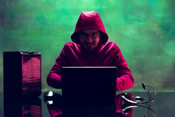 Photo of Portrait of man, computer geek, hacker working with laptop, breaking security system isolated over dark green background in neon
