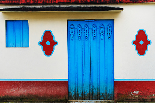 Front view of a white painted Colombian house with blue painted door and window and red painted patterns.