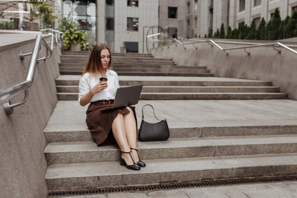 joven empresaria sosteniendo café y trabajando por computadora portátil en la calle de la ciudad - skirt brown fotografías e imágenes de stock