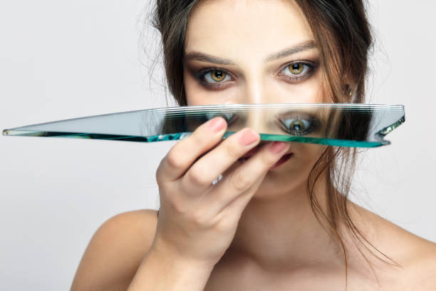 Portrait of a beautiful girl with a mirror splinter near eyes. Portrait of a beautiful girl with a mirror splinter near eyes. Female with mirror shard in hand posing on gray background. sliver of light on face stock pictures, royalty-free photos & images