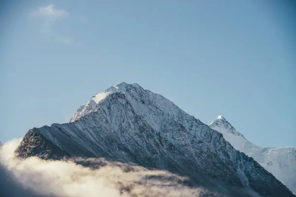 Beautiful view of snow-capped mountains above thick clouds in sunshine. Scenic bright mountain landscape with white-snow peak among dense low clouds in blue sky. Wonderful scenery with snowy pinnacle.