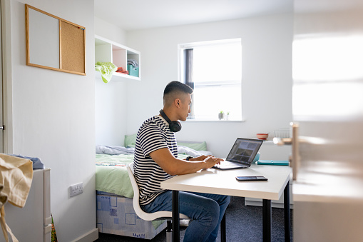 Male uni student sitting in his dorm bedroom while studying, using his laptop wearing headphones around his neck. He is in the North East of England.