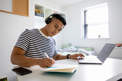 Male uni student sitting in his dorm bedroom while studying, using his laptop wearing headphones. He is writing in a notebook in the North East of England.