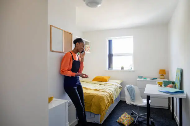 Female student standing talking on her mobile phone in her university bedroom in the North East of England. She is leaning against the wall smiling.