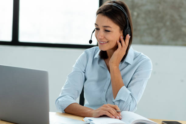 istock Woman customer support hotline operator with headset consults online client. Portrait beautiful caucasian young female call center worker sitting at the table in the office looking at laptop screen, smiles friendly 1369314800