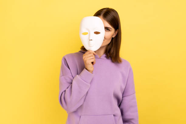 Woman removing white mask from face showing his smiling expression, good mood. Woman with dark hair removing white mask from face showing his smiling expression, good mood, pretending to be another person, wearing purple hoodie. Indoor studio shot isolated on yellow background. hypocrisy stock pictures, royalty-free photos & images