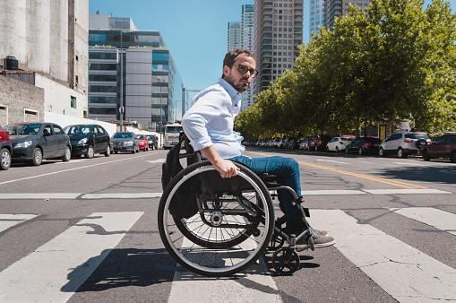 Caucasian Latino Hispanic adult business man, in a wheelchair attentively crossing the street with caution, in the middle of the avenue on the pedestrian stripe.