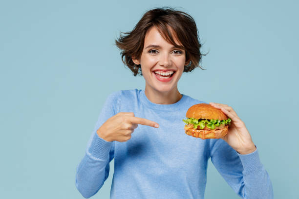 Young smiling fun caucasian fun cool woman 20s wearing casual sweater point finger on burger eating isolated on plain pastel light blue background studio portrait. People lifestyle junk food concept joven sonriente diversión caucásica diversión mujer cool de 20 años con suéter casual con el dedo acusador en hamburguesa comiendo aislado en un retrato de estudio de fondo azul claro pastel liso. concepto de comida chatarra de estilo de vida de las p