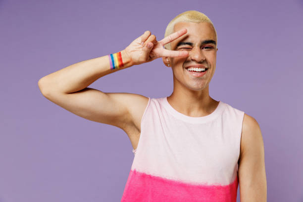 joven divertido y amigable hombre gay latino de 20 años con maquillaje que usa un ojo de cubierta superior rosa brillante con signo de victoria aislado en un fondo púrpura pastel simple retrato de estudio estilo de vida de las personas moda lgbtq concept - homosexual rainbow gay pride flag flag fotografías e imágenes de stock