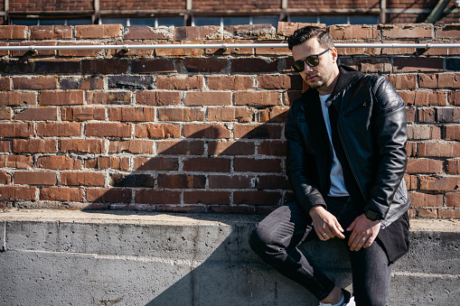 Young man wearing black jeans, a black leather jacket and sunglasses sitting in front of a brick wall deep in thought