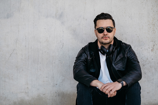 Man wearing wireless headphones, a smartwatch, sunglasses and a leather jacket sitting in front of a gray concrete wall