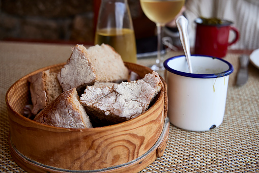 Various pieces of bread in a wooden bread basket. In the background two cups of broth and a glass of wine