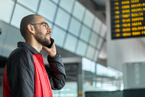 Young male speaking on smartphone by airport panel waiting for flight