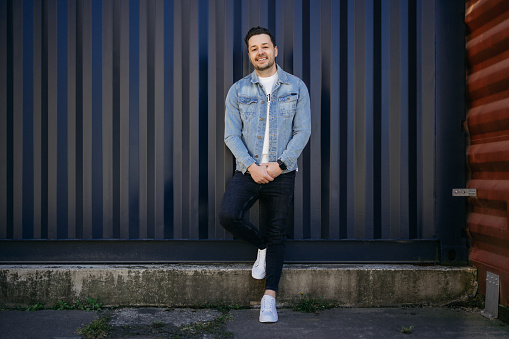 Young man wearing a casual denim shirt smiling in front of a blue shipping container