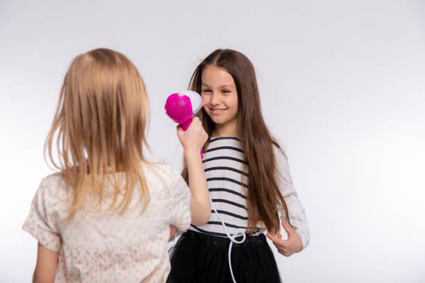 deux belles filles de 6 à 8 ans jouent avec le sèche-cheveux. les filles s’amusent à passer du temps ensemble et à sécher les cheveux sur fond blanc - female 8 9 years child excitement photos et images de collection