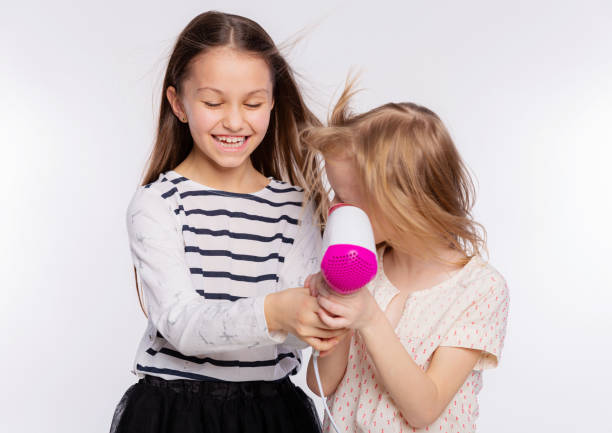 deux belles filles de 6 à 8 ans jouent avec le sèche-cheveux. les filles s’amusent à passer du temps ensemble et à sécher les cheveux sur fond blanc - female 8 9 years child excitement photos et images de collection