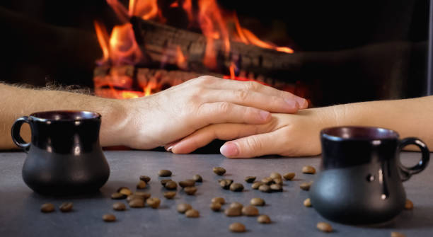 istock Romantic coffee break. A couple drinks coffee from ceramic coffee cups against a backdrop of burning fire and coffee beans. Close-up on hands. 1369337549