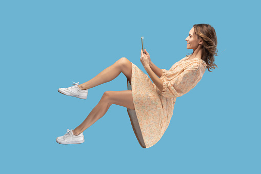 Hovering in air. Smiling girl in yellow dress levitating with mobile phone, reading message chatting happy joyful in social network online, surfing web while flying. indoor shot isolated on blue