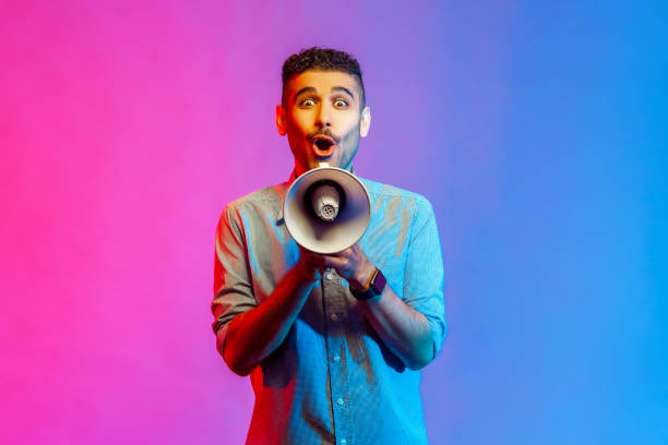 Man in shirt holding megaphone in hands and looking at camera with shocked facial expression. stock photo